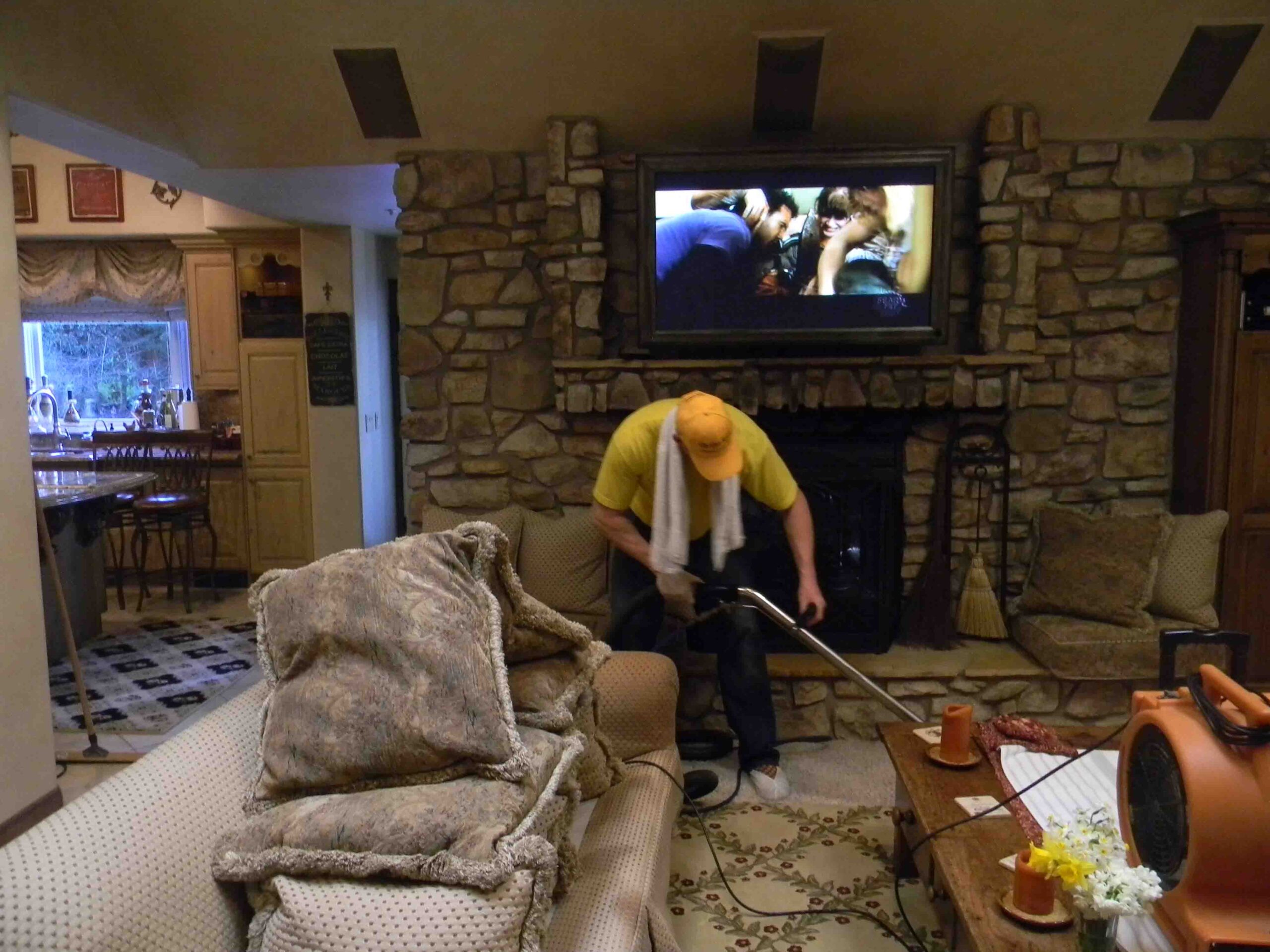 On-The-Spot Cleaning Services - Tony, owner, cleaning the living room of a mansion. His turbo-dryers are drying the couch and couch cushions, and he is steam-cleaning the carpets. Music videos are playing on the flatscreen on the wall.